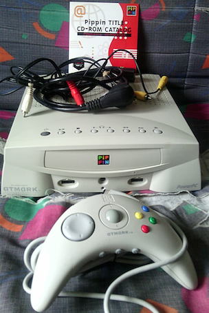 Console Pippin avec manette et câbles, étiquette de jeu CD-ROM "Pippin TITLE CD-ROM CATALOG" posée dessus